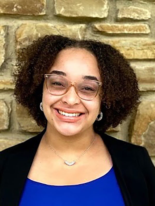 A headshot of Ayawna Kemp, who wears a blue shirt and dark blazer, and poses against a stone wall.