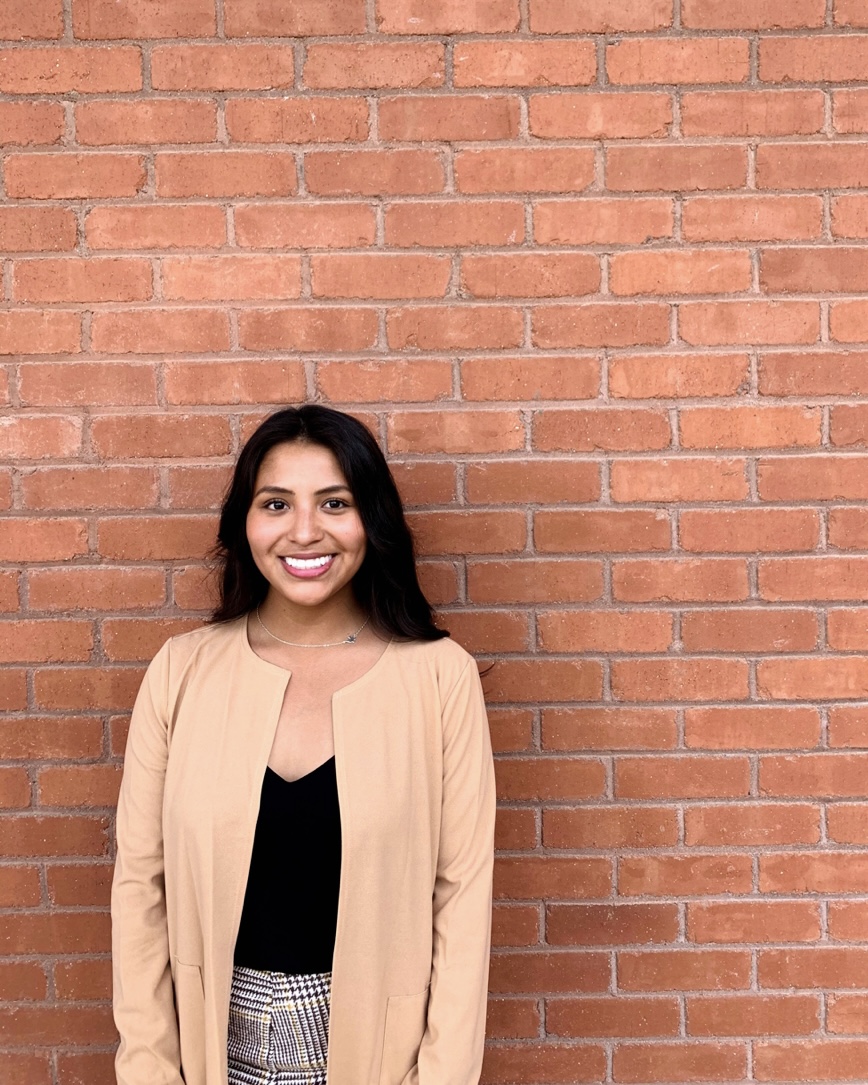 A headshot of Leslie Del Carpio, who wears a beige jacket and black shirt, and poses against a brick wall.