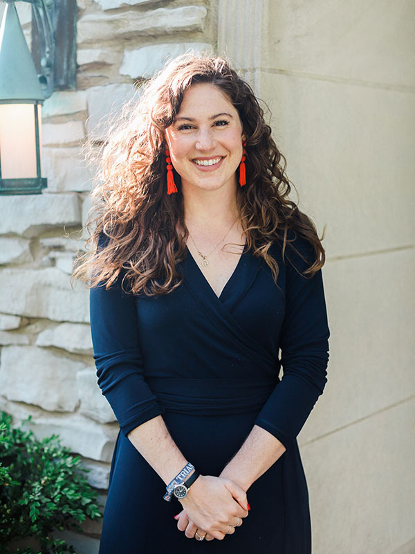 A headshot of Mackenzie Coulter-Kern, who wears a dark blue dress and poses outside against a limestone wall.