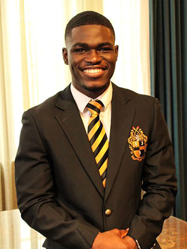 A headshot of Mout-Maine Moustapha, who wears a dark suit and yellow-and-black tie, and poses in front of a sunlit curtain.