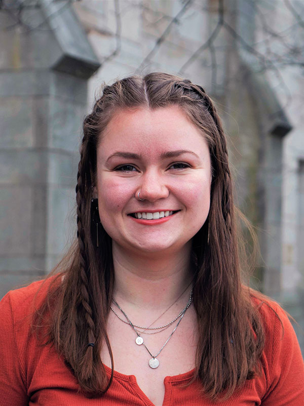 A headshot of Nora West, who wears an orange-red shirt and poses against a limestone building.