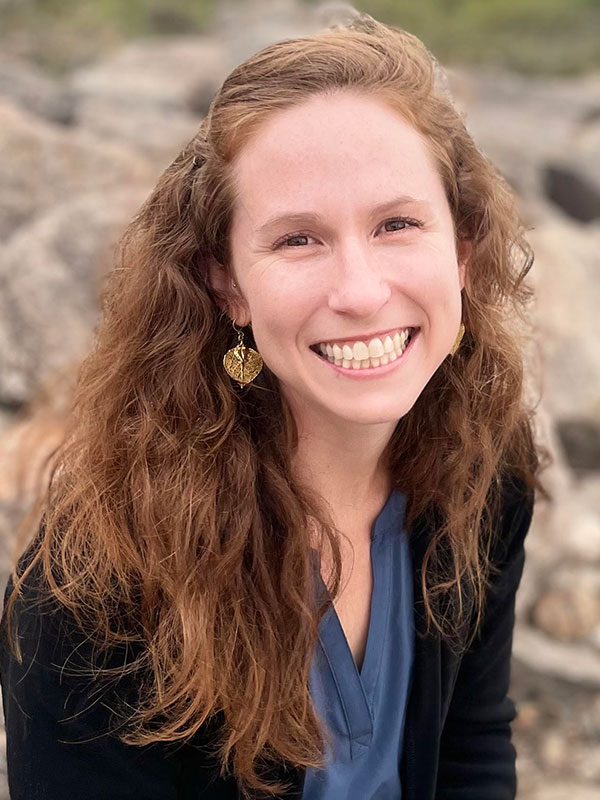 A headshot of Sarah Renkert, who wears a blue shirt and black blazer, and poses against a blurry, outdoor background.