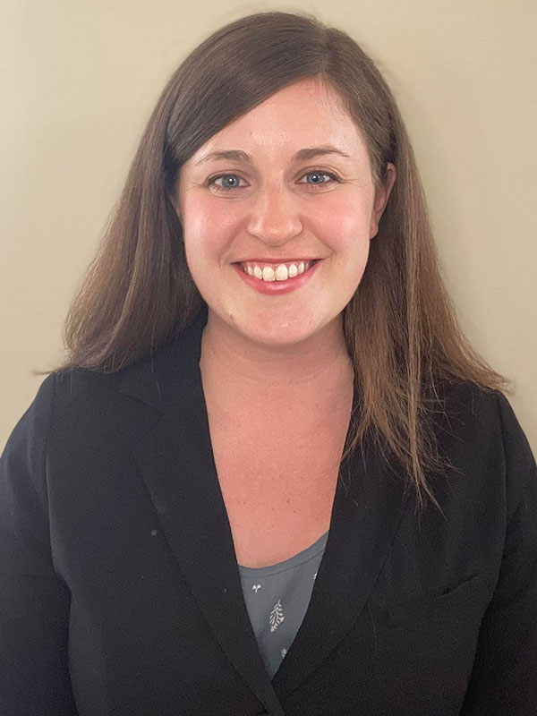 A headshot of Sarah TeKolste, who wears a black blazer and poses against a beige wall.