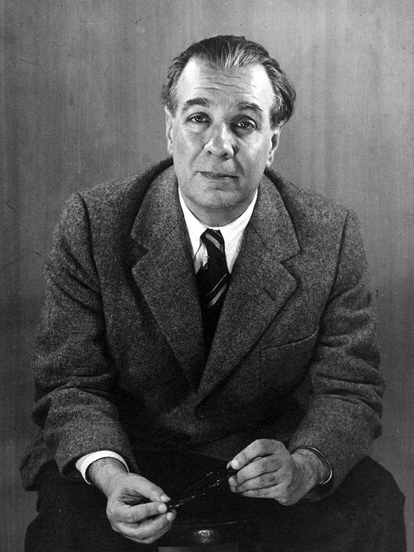 A black-and-white photograph of author Jorge Luis Borges, who wears a dark gray suit and holds his glasses in his hands.