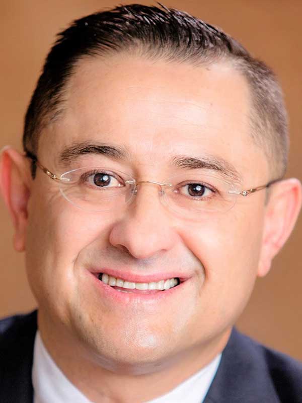 A headshot of César Félix-Brasdefer, who wears a dark suit and poses against a beige background.