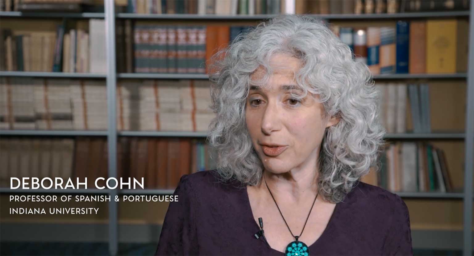 A screenshot of Deborah Cohn in the film, who's seated in front of a bookshelf.