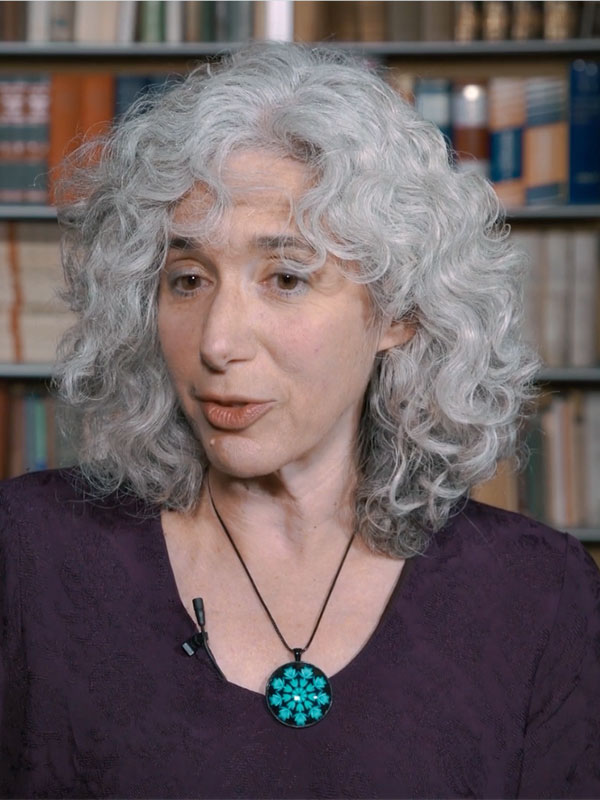 A headshot of Deborah Cohn, who wears a burgundy shirt and poses in front of a crowded bookshelf.