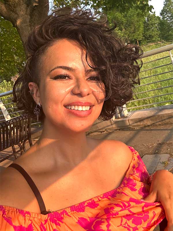 A headshot of Lais Lara Vanin, who wears a orange-and-red shirt and poses on a patio outside.