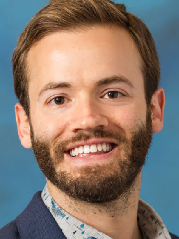 A headshot of Matthew Kanwit, who wears a blue blazer and poses against a blue background.