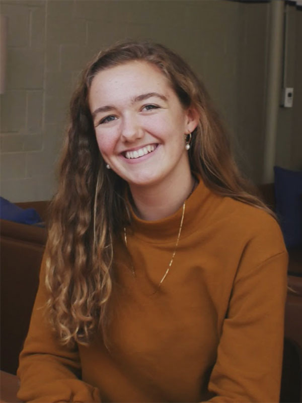 A headshot of Rose Schnabel, who poses in a dark orange sweater with a beige limestone wall in the background.