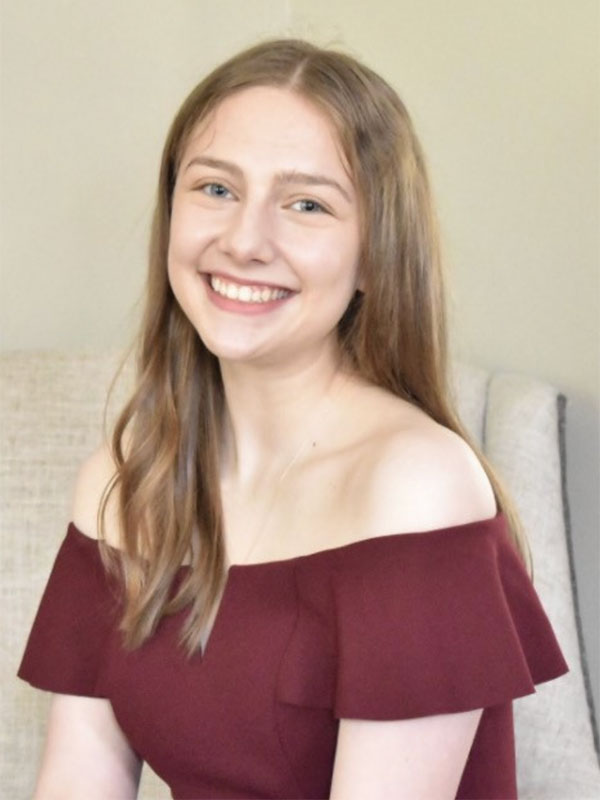 A headshot of Sarah Waters, who wears a dark red dress and poses seated on a beige chair.