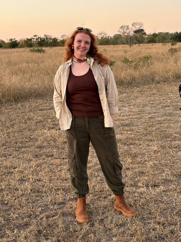Person standing outdoors in a grassy field at sunset, dressed in outdoor attire and smiling at the camera.