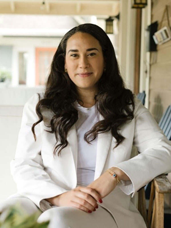 Person in a white suit sitting on a porch chair and smiling at the camera.