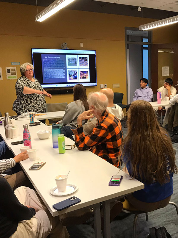 A person giving a presentation to a seated audience in a classroom with a large screen displaying text and images.