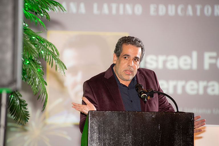 Person speaking at a podium during an event, with a projected background behind them.