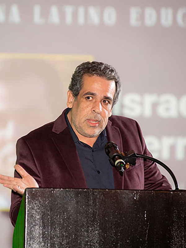 A person speaking at a podium during an event, gesturing with one hand in front of a projected background.