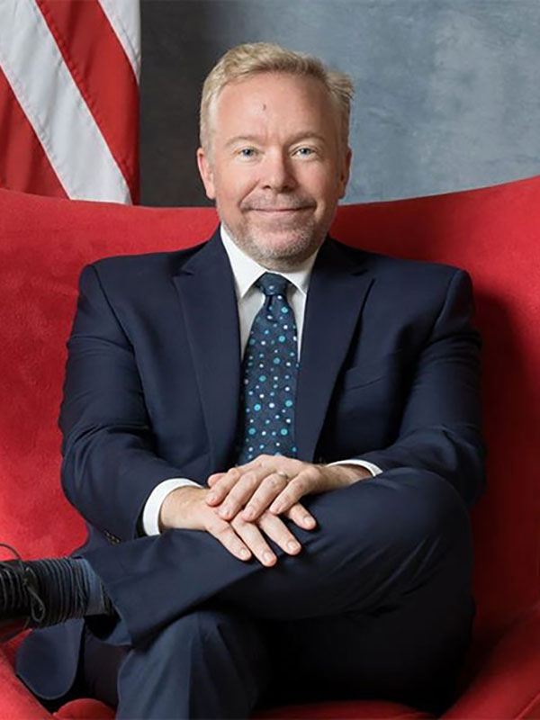 Person seated cross-legged in a red chair wearing a dark blue suit and tie, smiling with an American flag behind them.