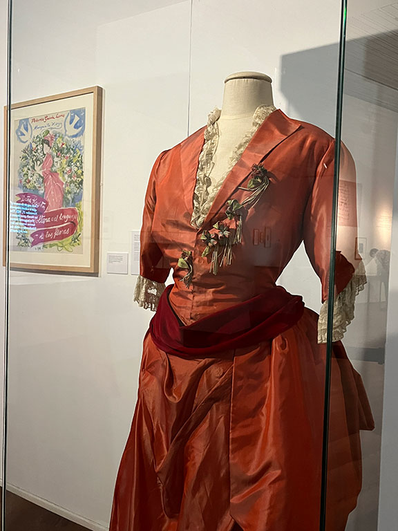 A bright orange and burgundy period dress with lace trim displayed in a glass case beside a colorful vintage poster.