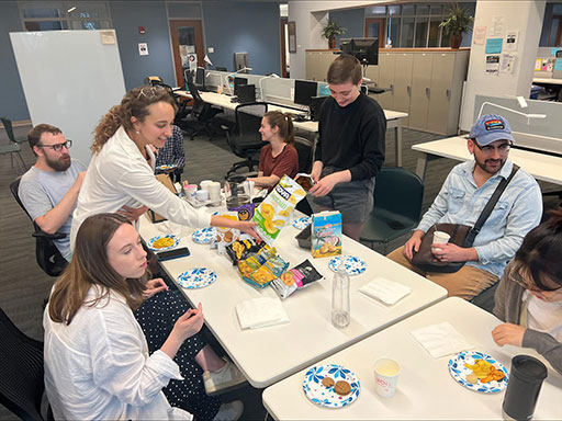 Group of people gathered around a table in an office setting, sharing snacks and talking casually.