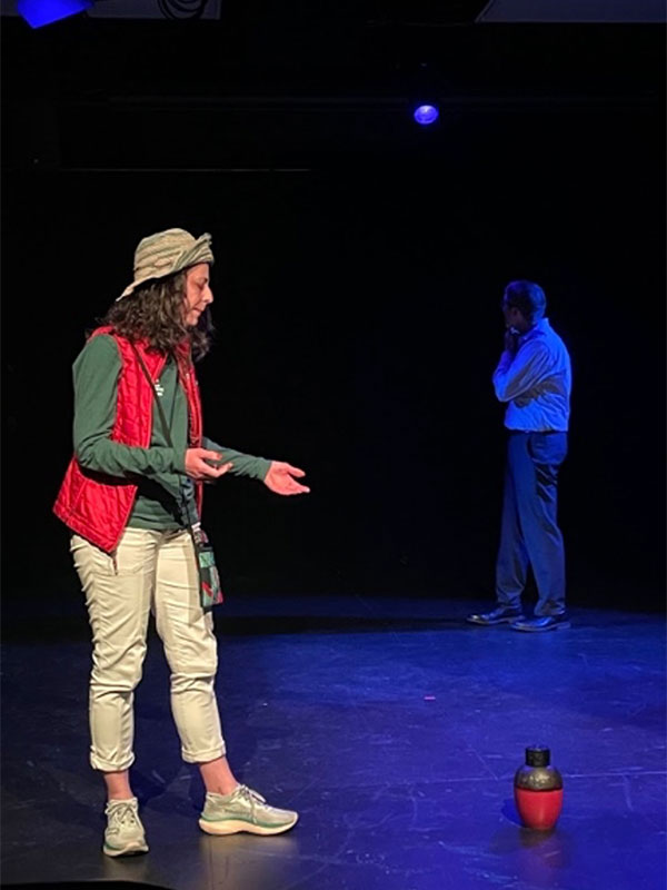 Two actors performing on a dimly lit stage, one speaking and gesturing while the other stands in the background.